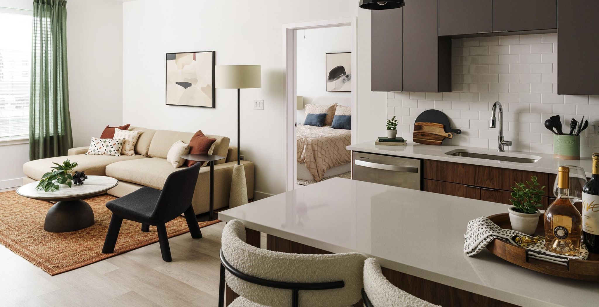 Modern open-plan living area with a beige sofa, black chairs, kitchen island, and a glimpse of a bedroom. Art on walls.