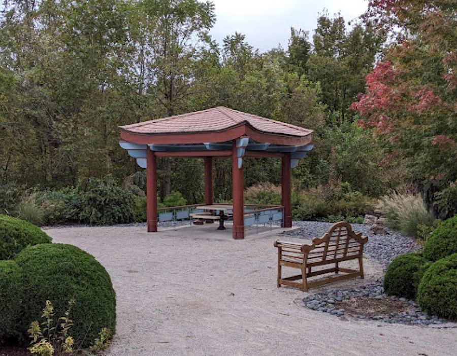 The Oslo A gazebo nestled in a park, encircled by lush trees and vibrant bushes, creating a serene outdoor space.

