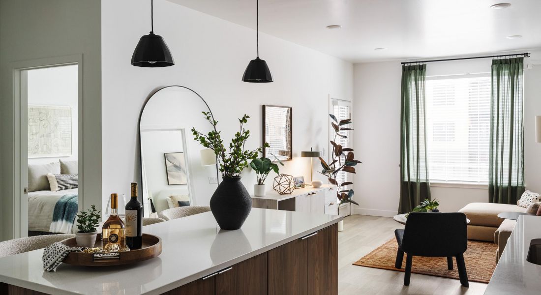 Modern kitchen interior with a wooden island, black pendant lights, and a cozy sitting area with plants and artwork.