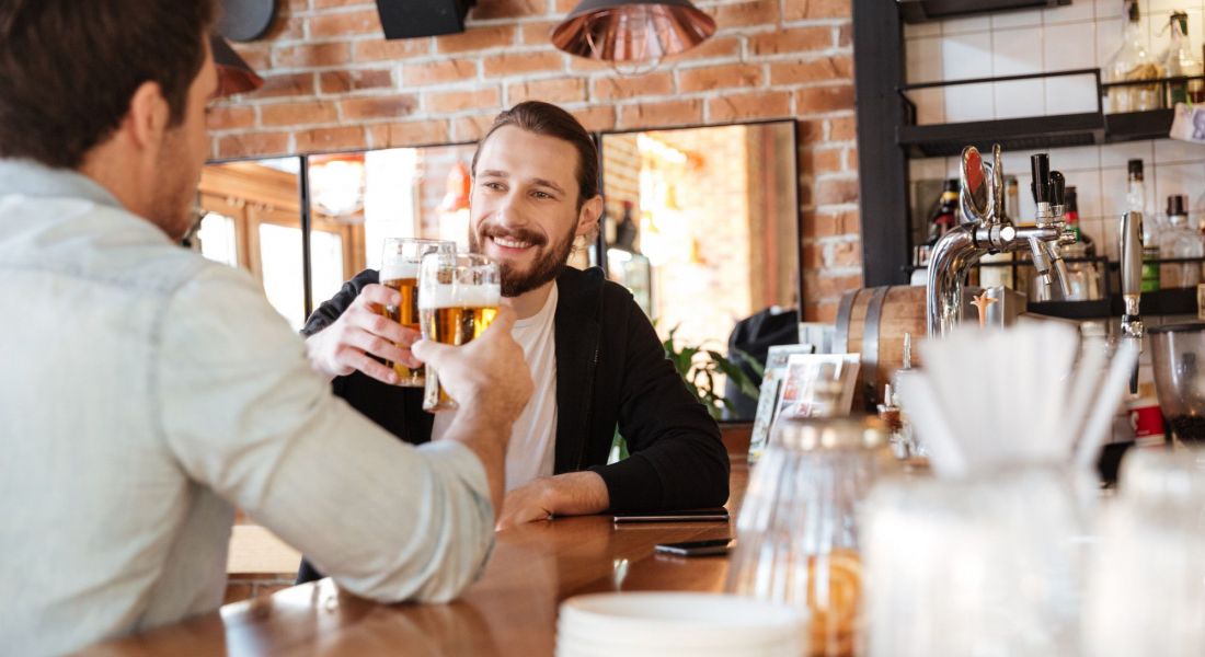 The Oslo Two men at a bar, one raising a beer in a toast, celebrating their time together.
