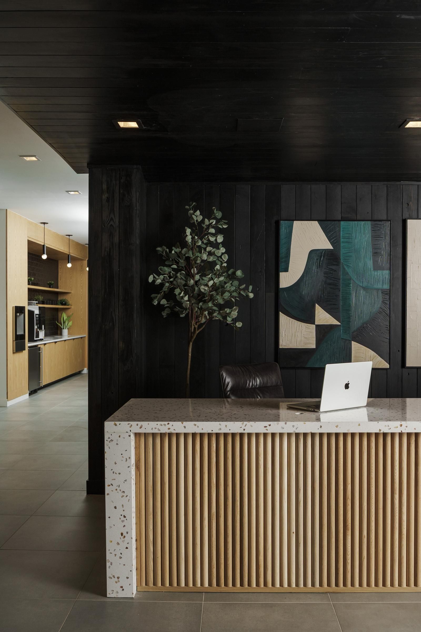 Modern reception area with a light wood desk, laptop, abstract art, and a tall plant. Room extends to a kitchen in the back.