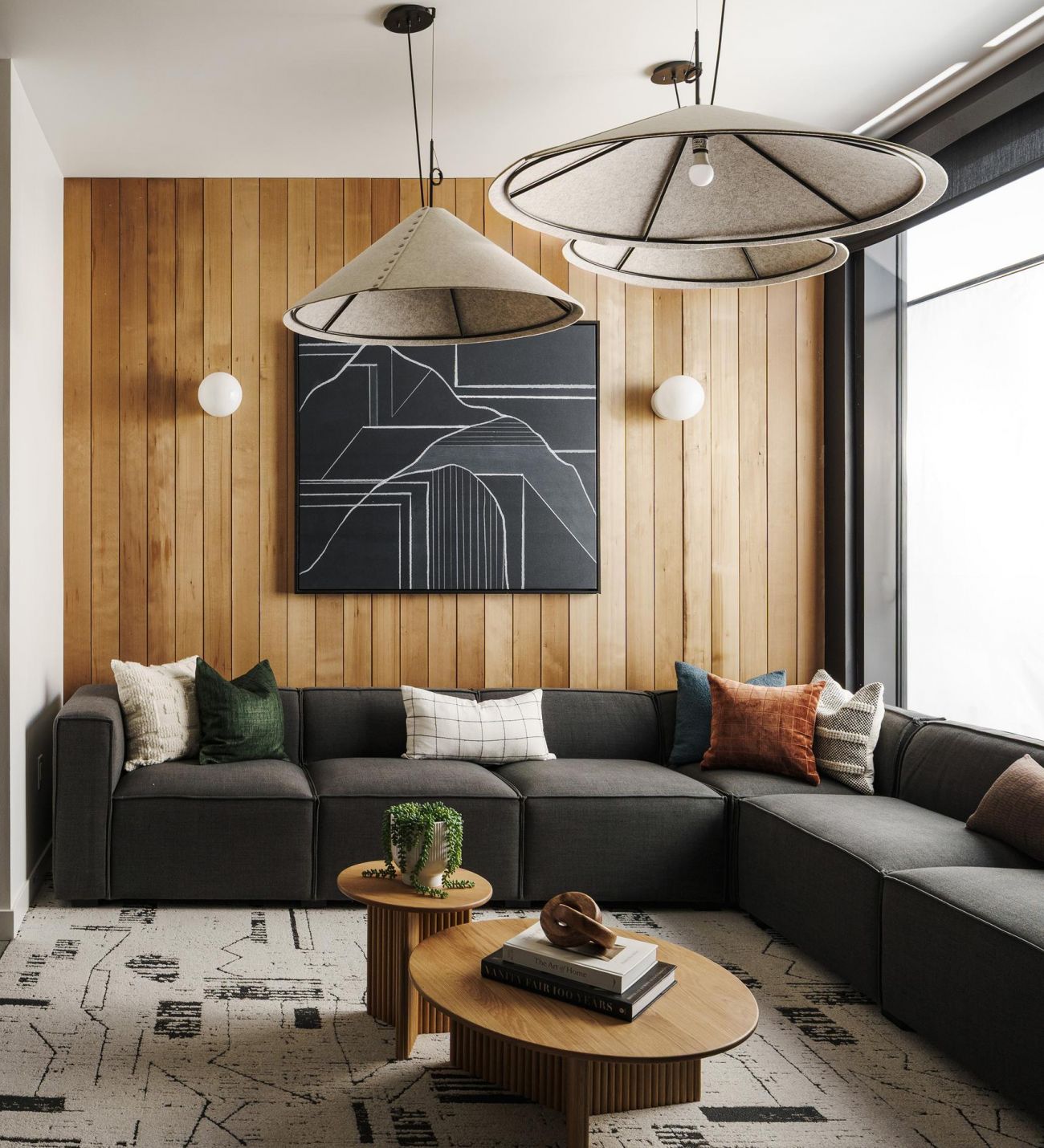 Modern living room with a dark sectional sofa, wooden wall, large window, and abstract art above small round tables.