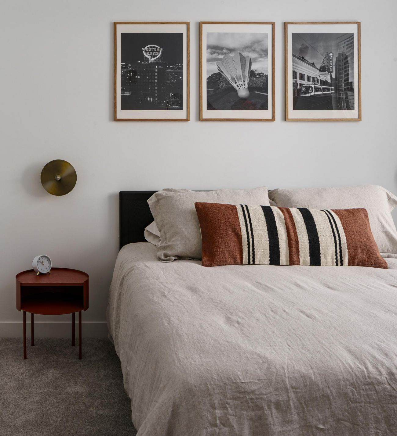 Modern bedroom with a beige bed, striped pillows, red side table, and three black-and-white photos on the wall.