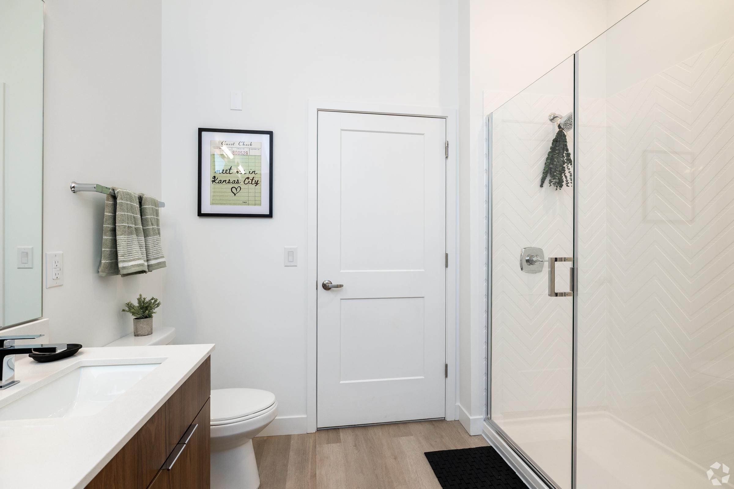 The Oslo with glass shower, white walls, wood vanity, and green towels hanging beside framed wall art.